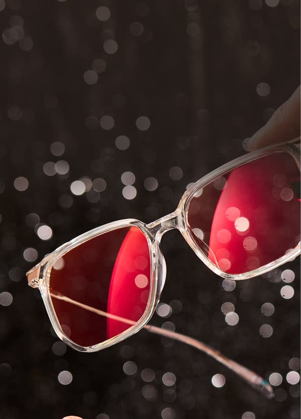 EyeQLenz with Zenni ID Guard eyeglasses with pink-tinted lenses held against sparkling holiday lights background.