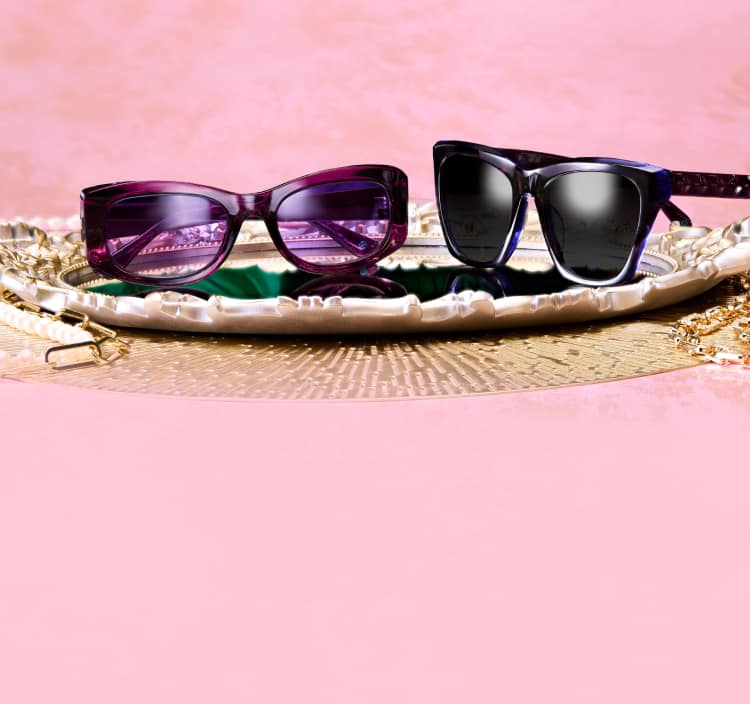 Two pairs of Zenni glasses from the Iris Apfel collection, one purple and one black, on a decorative tray with a pink background.