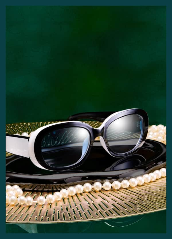 Black and white Zenni glasses from the Iris Apfel collection sit on a pearl-accented tray with a forest green background.