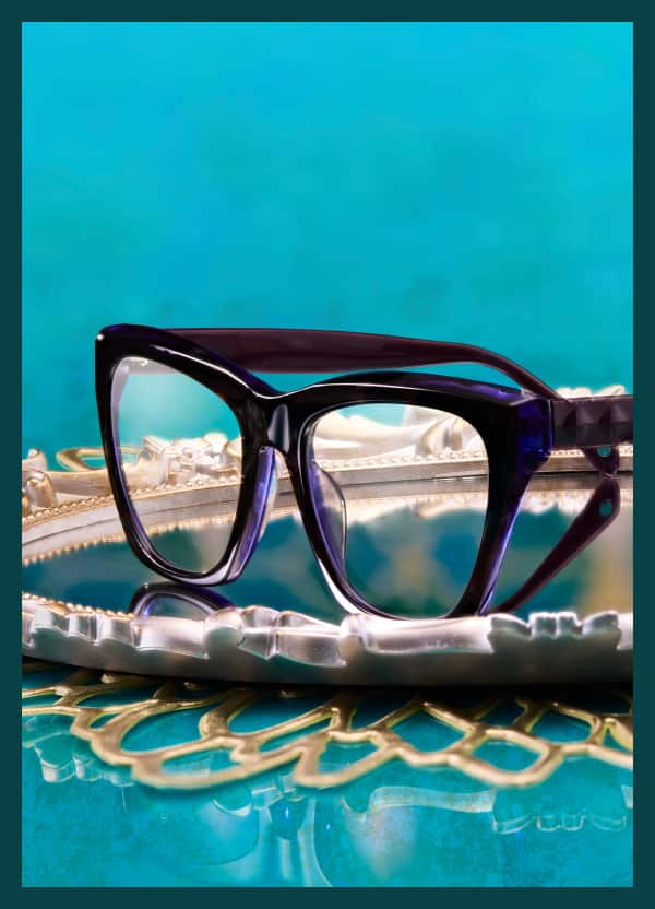 Purple Zenni glasses from the Iris Apfel collection sit on a mirrored tray with a turquoise background.