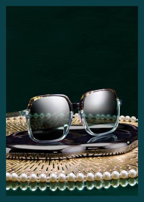 Square Zenni glasses from the Iris Apfel collection sit on a gold tray with pearl accents and a dark green background.