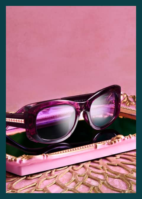 Purple Zenni glasses from the Iris Apfel collection sit on a pink-framed tray with a pink background.