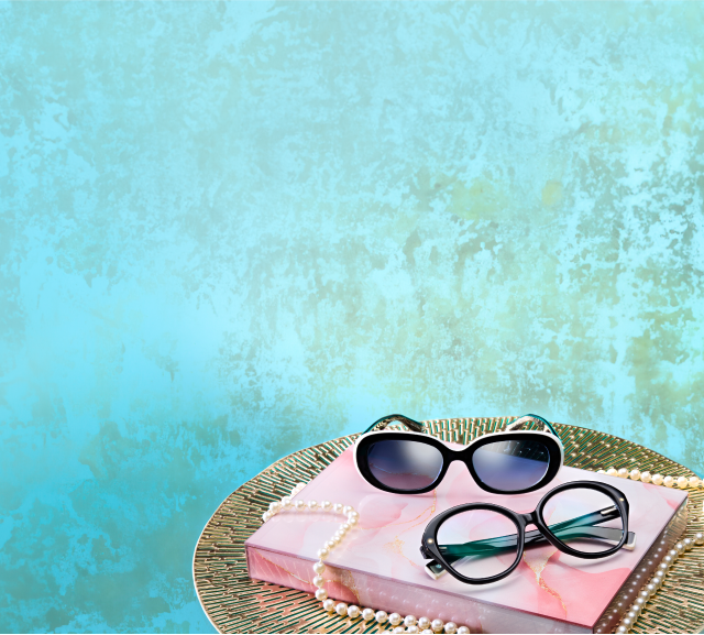 Two pairs of Zenni glasses from the Iris Apfel collection, one black and white and one black with green arms, sit on a pastel pink book and gold tray with a string of pearls.