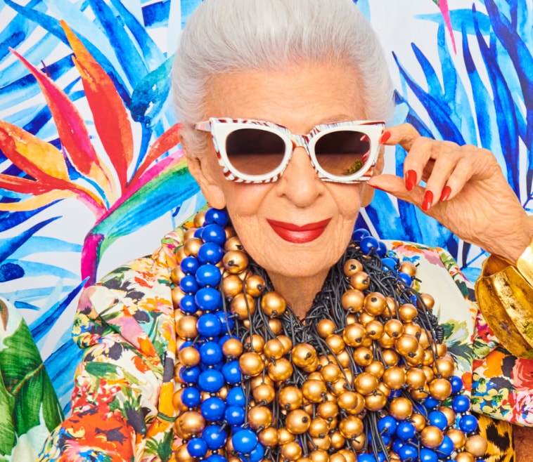 Iris Apfel wearing white cat-eye frames with a red and light blue patterned rim. 