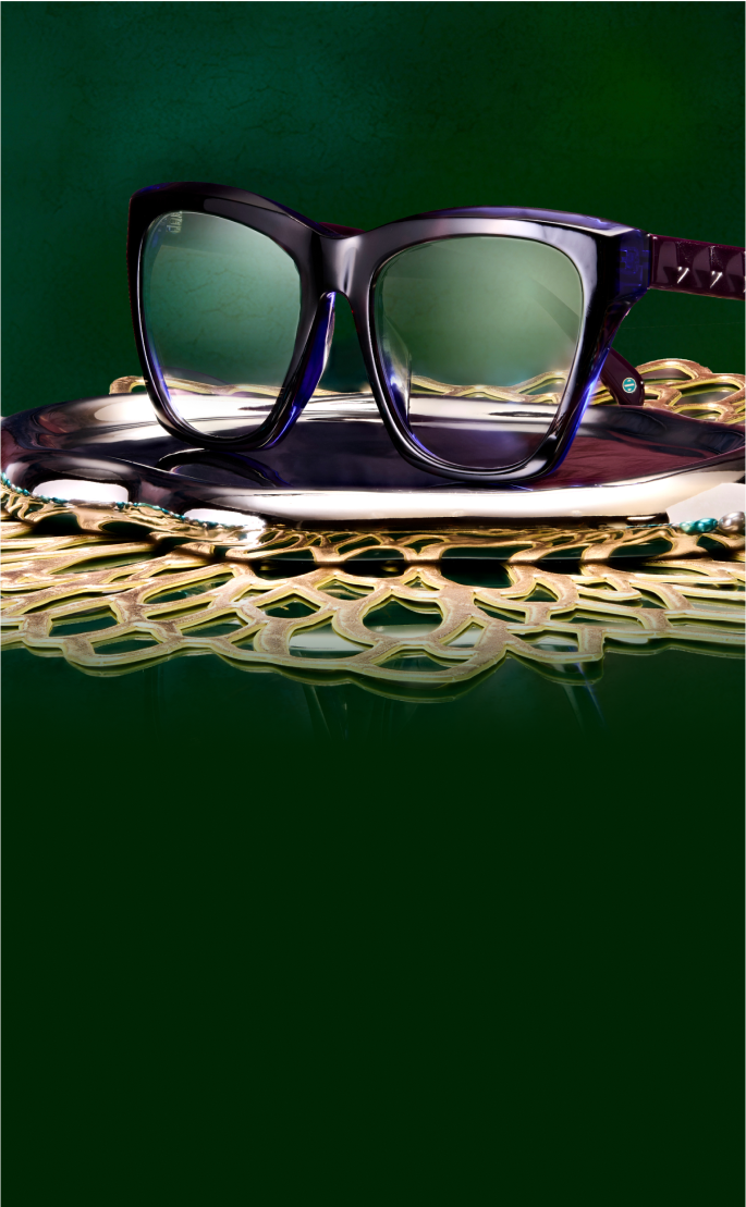 A pair of dark-framed glasses 'Iconic Iris' is displayed on a decorative gold tray against a rich green background. A strand of turquoise and white beads is draped casually on the tray beside the glasses.