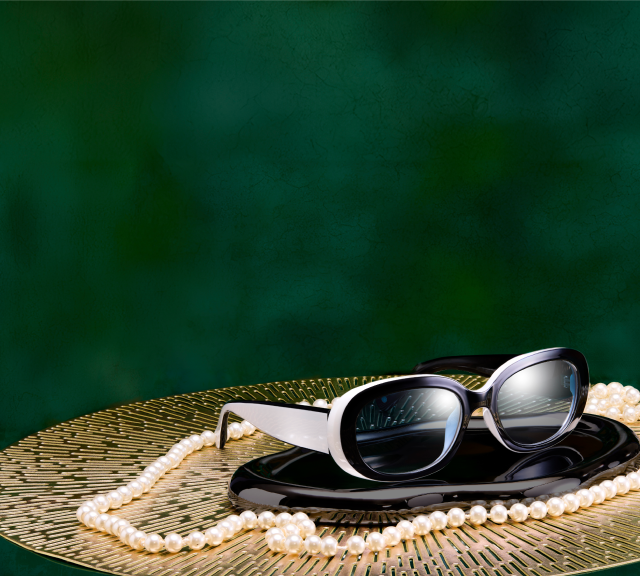 A image featuring a pair of eyeglasses with round black frames, resting on on a decorative tray adorned with a pearl necklace, with a textured dark green background.