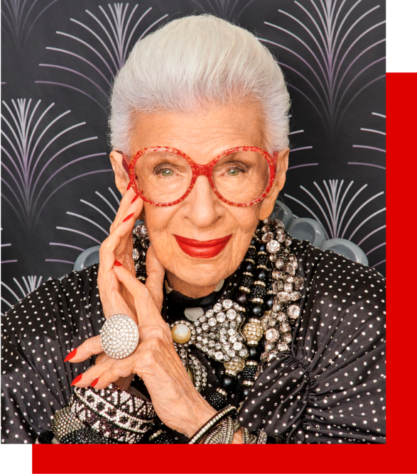 Image of Iris Apfel wearing Zenni Iris Apfel round glasses #4452518. She is wearing bright red lipstick and is in front of a black patterned wall.