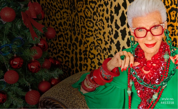 Iris Apfel wearing a brightly colored outfit, with Zenni glasses and eyewear chain in front of a leopard-print wall.