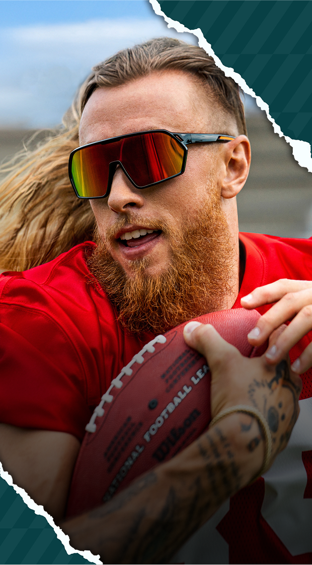 George Kittle wearing mirrored sport sunglasses and a red jersey while holding a football on the field.