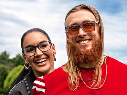 George Kittle and Claire Kittle smile outdoors under a partly cloudy sky. Claire wears rectangular eyeglasses and hoop earrings, while George wears red-tinted sunglasses, a red jersey, and gold chains.