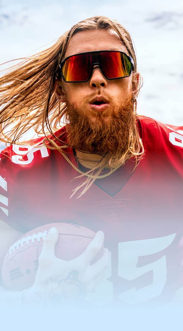 George Kittle in a red jersey and mirrored sport sunglasses, running on a football field while holding a football.