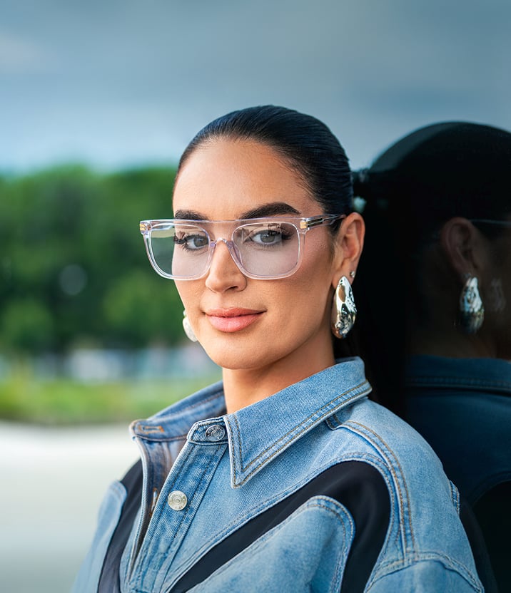 Claire Kittle in Zenni clear and blue “Defender” square glasses that have blue tinted lenses, with her hair slicked back into a ponytail wearing a chic denim dress and chunky earrings.
