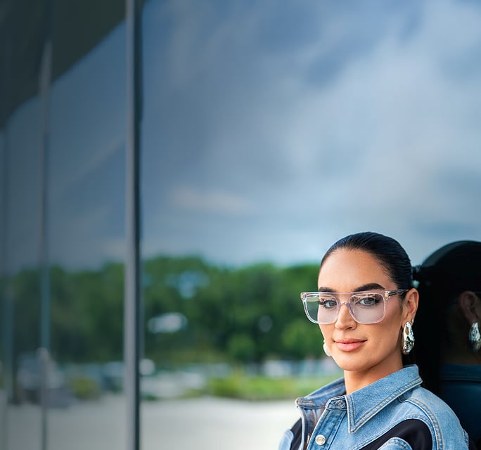 A portrait of Claire Kittle wearing oversized, clear-framed glasses and a denim jacket against a soft-focus outdoor background.