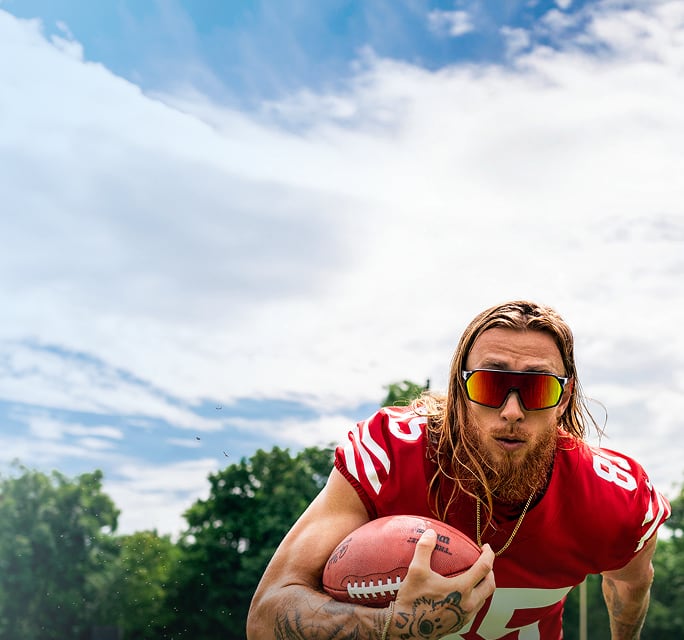 George Kittle running with a football in a 49ers jersey while wearing large, wraparound performance sunglasses with red-orange mirrored lenses.