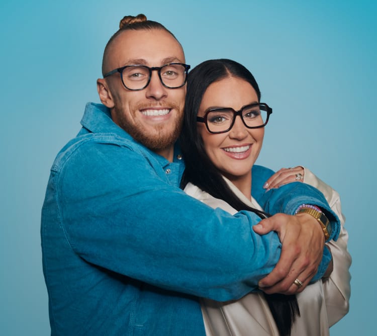 George Kittle and wife Claire, wearing Zenni glasses, smiling and embracing against a light blue background.