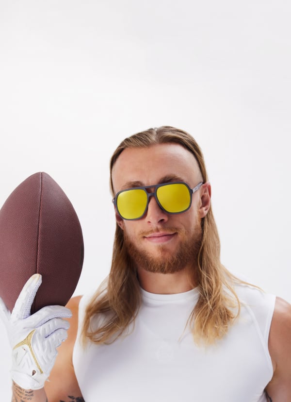 George Kittle, 49er, with long hair, holding a football, wearing yellow lens sunglasses and white tank, white background. (STYLE NAME & #1144812).
