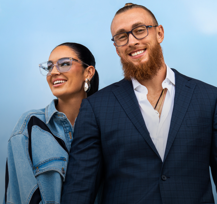 San Francisco 49ers tight end George Kittle is wearing blue Zenni “Hawk Vision” glasses that have clear optical lenses and his wife, fashionista Claire Kittle is wearing clear Zenni “Defender”glasses, holding hands and smiling in stylish outfits against a blue sky.