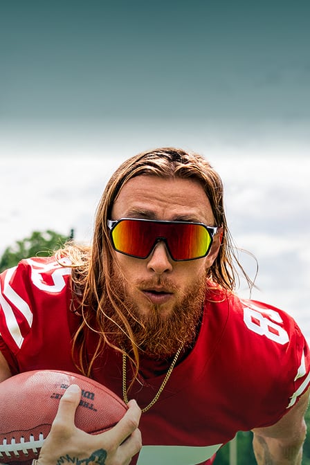 George Kittle in his football jersey and Zenni mirrored sunglasses holding a football.