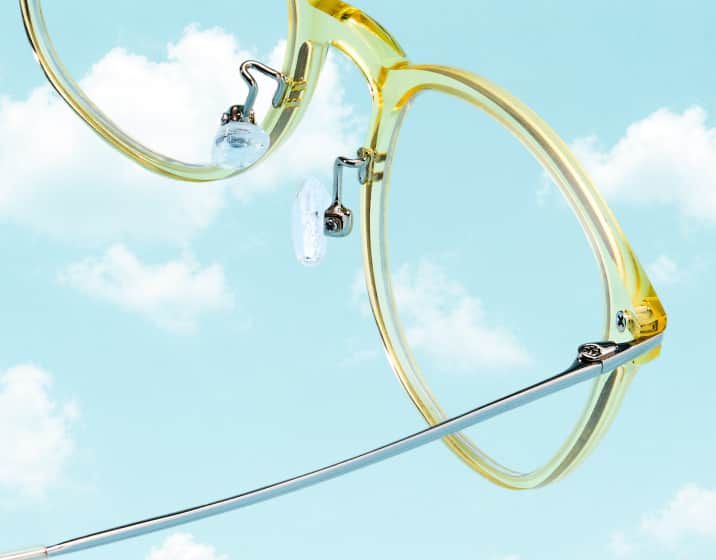 Close-up of translucent yellow round eyeglasses with thin metal temple arms, floating against a bright blue sky filled with soft white clouds.