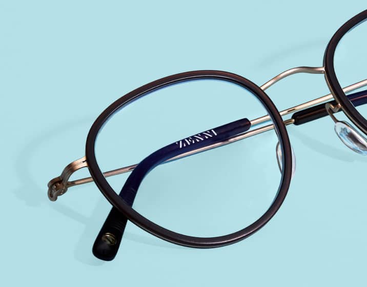 Close-up of dark brown round eyeglasses with a gold metal bridge and the Zenni logo printed on the inside of the temple arm, resting on a solid light blue background.
