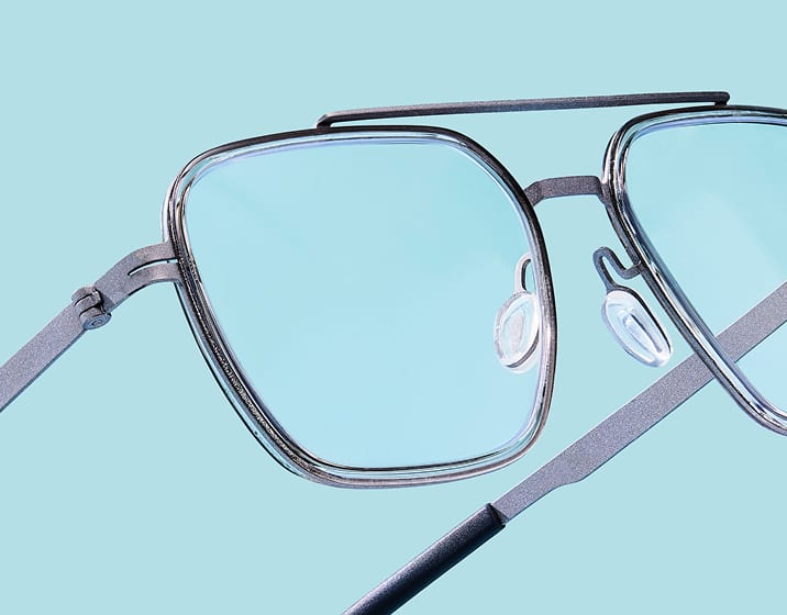 Close-up of grey geometric aviator-style eyeglasses with a double bridge and thin metal temples, floating on a solid light blue background.