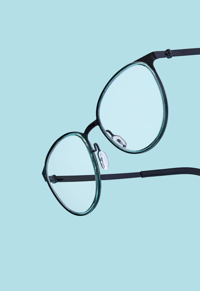 Round eyeglasses with thin dark green frames floating at an angle on a solid light blue background.