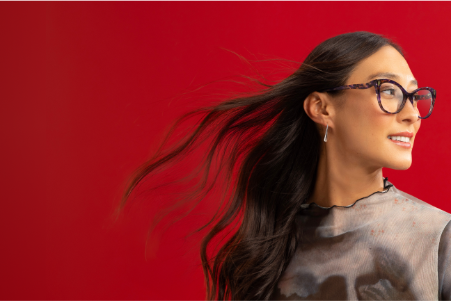 Woman with flowing dark hair wears purple tortoiseshell glasses against a bold red background featuring a subtle snake illustration.