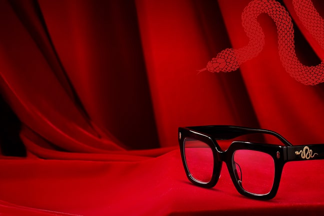 A pair of black glasses with gold snake detailing rests on luxurious red fabric. A stylized red snake illustration slithers in the background, complementing the bold, dramatic setting.