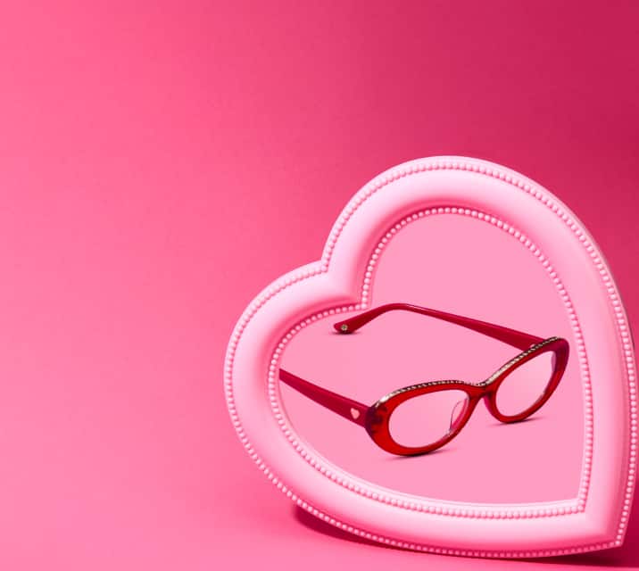 A pair of red oval glasses with gold heart accents on the arms centered inside a pink heart-shaped picture frame against a pink background