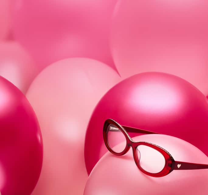 A pair of heart-shaped tortoiseshell glasses resting inside a pink heart-shaped frame against a solid pink background.'