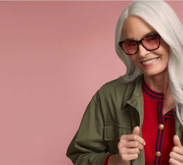 Woman with silver hair smiling, wearing a olive jacket and Zenni FL-41 rose-tinted glasses, against a pink background.