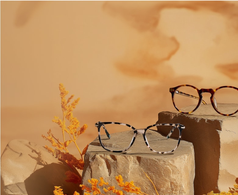 The image shows two pairs of stylish glasses displayed on stone-like pedestals against a warm, desert-inspired background.