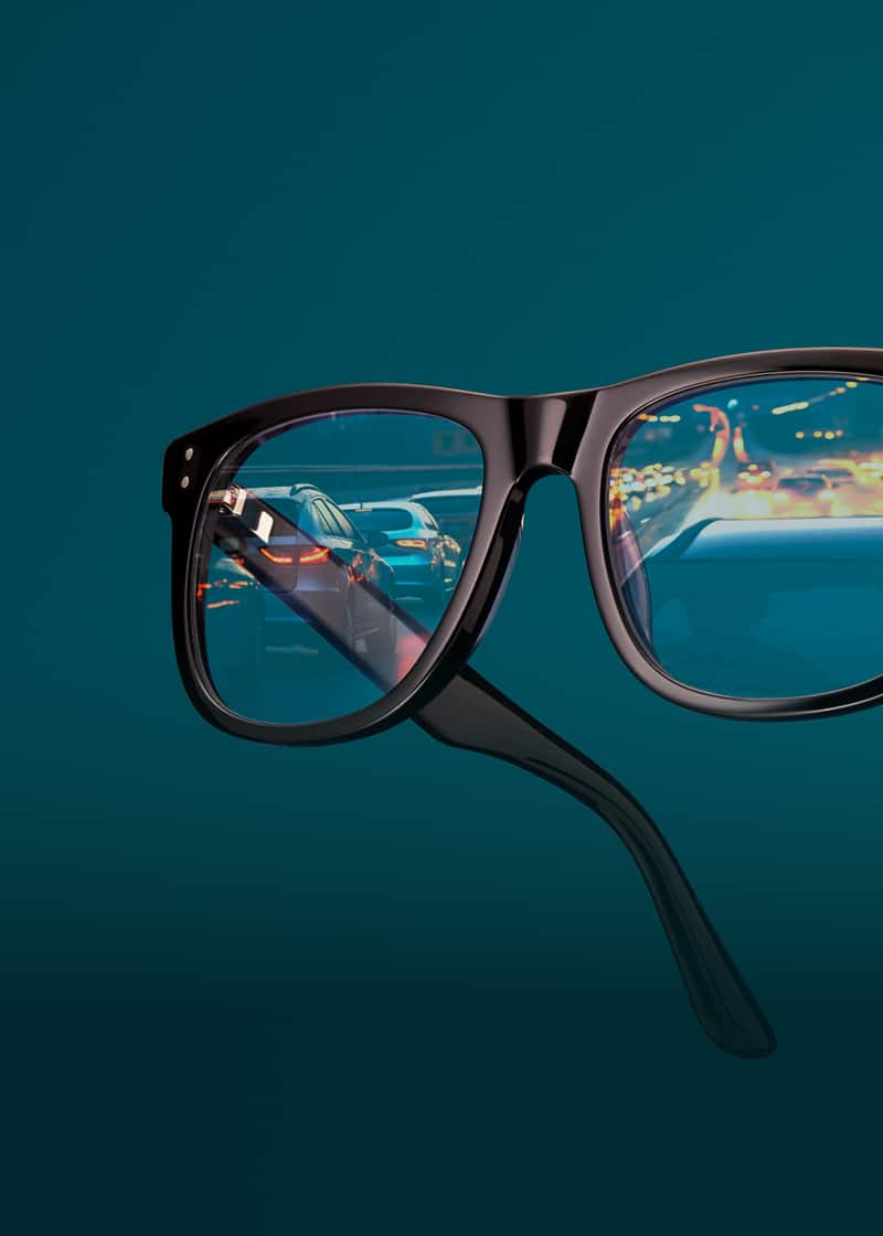 Square black glasses with nearly clear night driving lenses, reflecting a street at night. Background is dark teal.