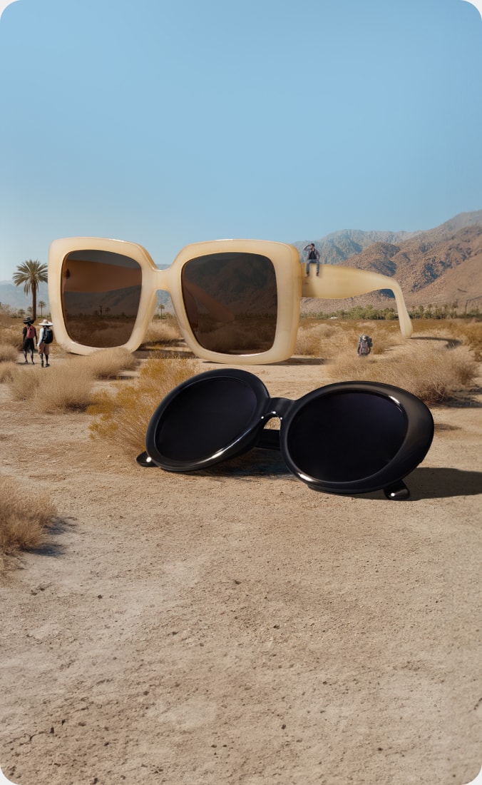 A desert scene features two oversized pairs of sunglasses placed among dry bushes and sandy terrain. One pair, with large square frames T01431515 in a beige color, stands upright with tiny people positioned on and around it. The other pair, with black round frames 2023021, lies on the ground.