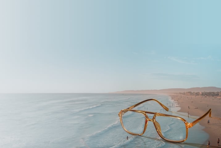 A striking pair of oversized amber-hued glasses floats over a sunlit coastline. The crystal-clear frame contrasts beautifully against the soft ocean waves and golden beach, merging retro style with relaxed summer vibes.