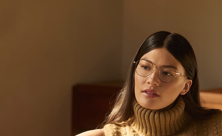 A woman wearing thin, metal-framed glasses sits in warm, soft lighting, dressed in a tan sleeveless turtleneck sweater.