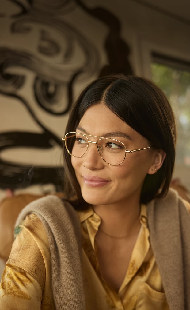 A woman with Zenni Progressive glasses sits contentedly, wearing a gold blouse under a beige cardigan, with abstract art in the background.