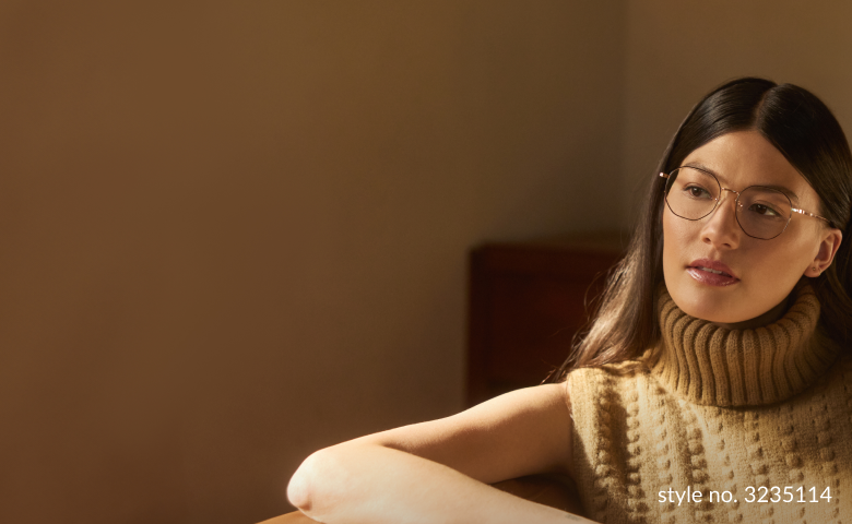 Thoughtful young woman with Zenni Progressive glasses looking away pensively, wearing a chunky turtleneck sweater, in a warmly-lit room.
