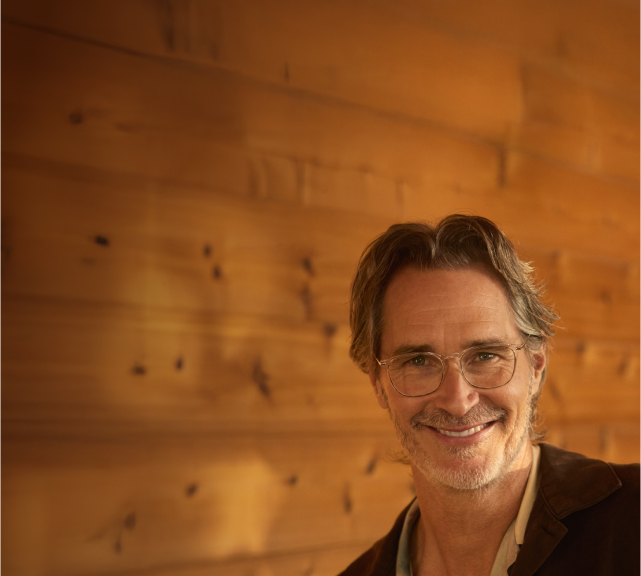 A man with graying hair and a beard, smiling warmly while wearing thin-framed glasses. He is standing in front of a wooden wall with soft lighting, creating a cozy and relaxed atmosphere.