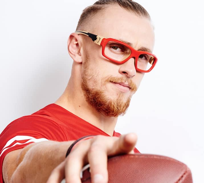George Kittle, San Francisco 49ers. Image of George Kittle wearing Zenni sport protective goggles #743528, holding a football pointed at the camera, against a white background.