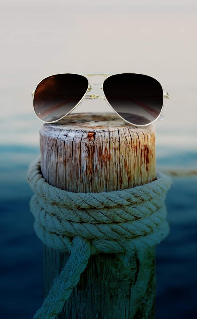 A pair of aviator sunglasses with dark lenses rests on a weathered wooden post by the water, with a blurred background of soft blue hues resembling the ocean or sky.