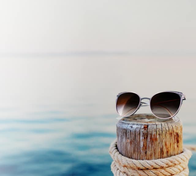 A pair of aviator sunglasses with dark lenses is resting on a weathered wooden post by the sea, with soft, blurred blue and white tones in the background, evoking a peaceful, beachside atmosphere.