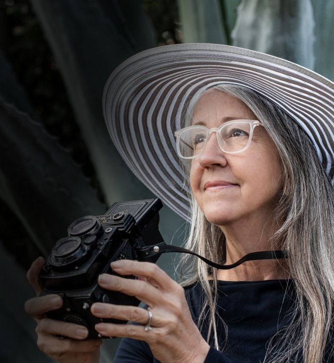 Image of a gray-haired woman wearing Zenni glasses and a striped large hat, holding a camera.