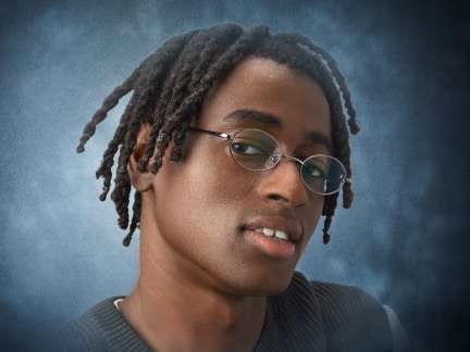 Close-up portrait of a person with twisted hair and glasses, smiling slightly against a textured, gradient blue background.