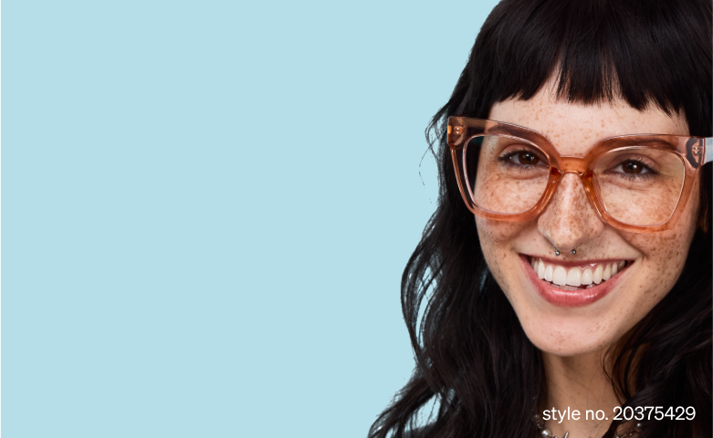 Woman with long dark hair and freckles, smiling brightly in oversized brown translucent glasses, against a pale blue background.