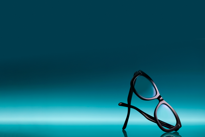 Stylish black eyeglasses with blue-reflective lenses standing upright on a glossy teal gradient background.