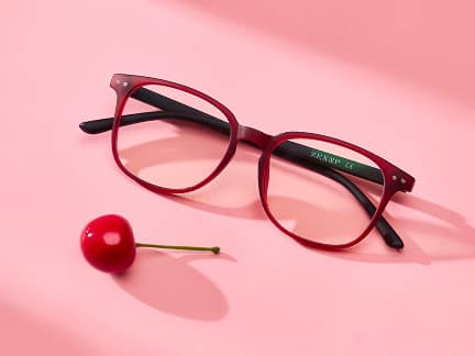 A pair of red eyeglasses with black arms lies on a pink surface surrounded by three cherries.