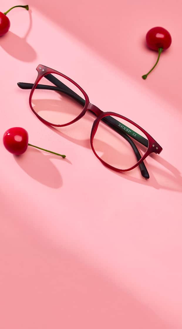 A pair of red eyeglasses with black arms lies on a pink surface surrounded by three cherries.