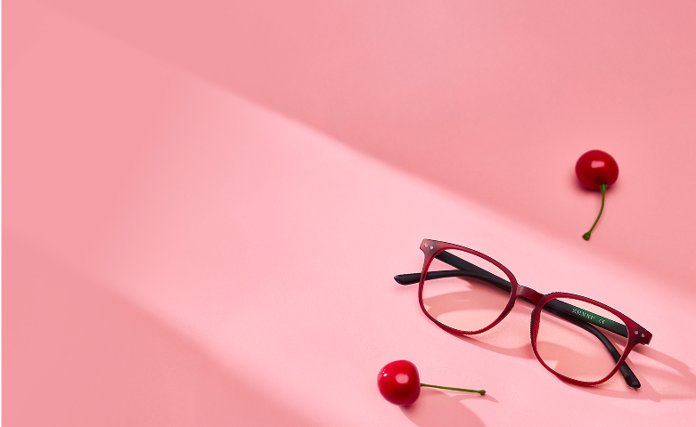 A pair of red eyeglasses with black arms lies on a pink surface surrounded by three cherries.
