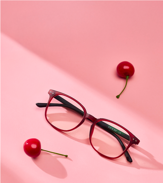 A pair of red eyeglasses with black arms lies on a pink surface surrounded by three cherries.